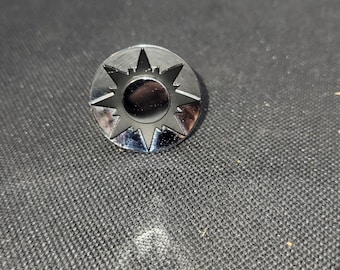 May include: A shiny, silver-colored metal pin with a sunburst design. The pin features a central circle surrounded by pointed rays, creating a star-like appearance. The pin is circular and has a reflective surface.