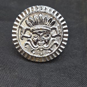 May include: A circular silver-toned metal button featuring a raised relief design of a stylized face wearing a feathered headdress. The button has a textured, ribbed outer border and a metallic finish with intricate detailing on the central figure.