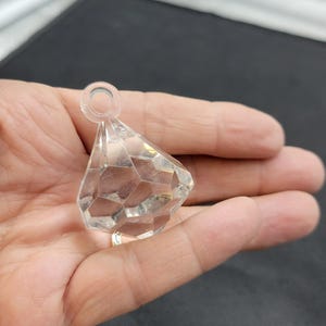 May include: A clear, faceted crystal pendant with a diamond shape. The pendant has a small loop at the top for hanging. The crystal is held in a hand against a dark background. The pendant is likely for jewelry or decorative purposes.