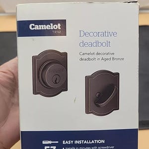 May include: A box for a Camelot Trim decorative deadbolt in Aged Bronze. The box shows a close-up of the deadbolt with a keyhole and a side view of the deadbolt with a handle. The box says "Easy Installation" and lists the steps: "Installs in minutes with screwdriver", "Guaranteed to fit on standard doors", "Self-guiding screws align and snap themselves into place". The box also says "Installation Videos at Schlage.com" and "Package Includes Everything You Need."