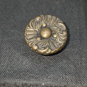 May include: A round, antique brass-colored knob with an ornate floral design. The knob features a central raised sphere surrounded by detailed leaf and swirl patterns. The metal has a slightly aged appearance, suggesting vintage or antique origins.