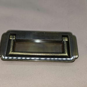 May include: A rectangular, antique-style drawer pull with a bronze finish. The pull has a curved handle and decorative edges. The hardware is designed for furniture and cabinets, offering a vintage aesthetic.