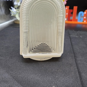 May include: A clear, rectangular glass light fixture with a white frame. The glass has a series of vertical lines that create a textured effect. The fixture is designed with an arched top and a rounded base.