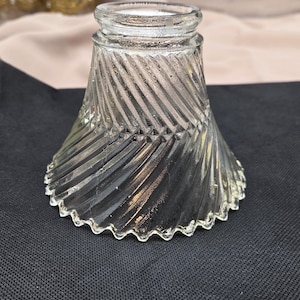 May include: Clear glass lampshade with a flared, conical shape. The shade features a textured, diagonal pattern and a scalloped edge. The lampshade is designed to diffuse light and add a decorative element to a lamp.