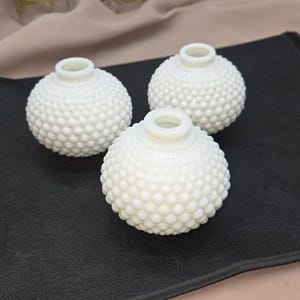 May include: Three white, round, hobnail-textured vases. The vases have a small opening at the top and are arranged on a black surface. The background is a neutral color.