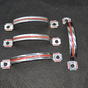 May include: Four curved metal drawer pulls with a polished chrome finish and a central red stripe. Each handle features two square mounting plates with pre-drilled holes at the ends, designed for attachment to furniture or cabinetry surfaces.