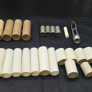 May include: An assortment of cylindrical paper tubes in various sizes and colors, including beige, tan, and gray. Also includes a metal and glass tool. The tubes are arranged on a black surface.
