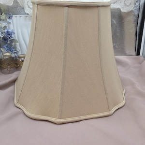 May include: A beige lampshade with a scalloped bottom edge. The lampshade has a slightly flared shape and is made of a textured fabric. The top and bottom edges are trimmed with a lighter beige material. The lampshade is a home decor item.