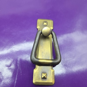 May include: A brass-toned cabinet pull with a rectangular backplate and a triangular handle. The backplate has square accents at the top and bottom. The handle is a dark, contrasting color. The pull is designed for furniture.