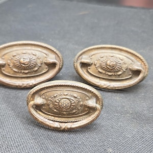 May include: Three oval-shaped metal drawer pulls with a bronze-toned finish. Each pull features a central decorative medallion with intricate floral-like detailing, a rope-textured border, and a hinged drop handle that rests against the base plate.
