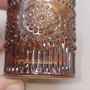 May include: Close-up of a brown glass tumbler with a detailed, embossed floral design. The glass has a cylindrical shape with vertical lines and small, raised dots. The design is repeated around the glass.