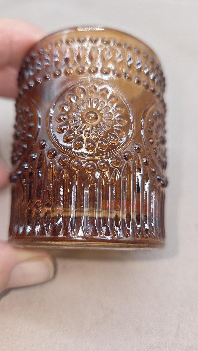 May include: Close-up of a brown glass tumbler with a detailed, embossed floral design. The glass has a cylindrical shape with vertical lines and small, raised dots. The design is repeated around the glass.