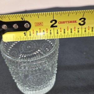 May include: A clear glass votive candle holder with a decorative pattern is measured with a yellow Craftsman tape measure. The tape measure reads approximately 2 inches across the top of the glass. The candle holder is on a dark surface.