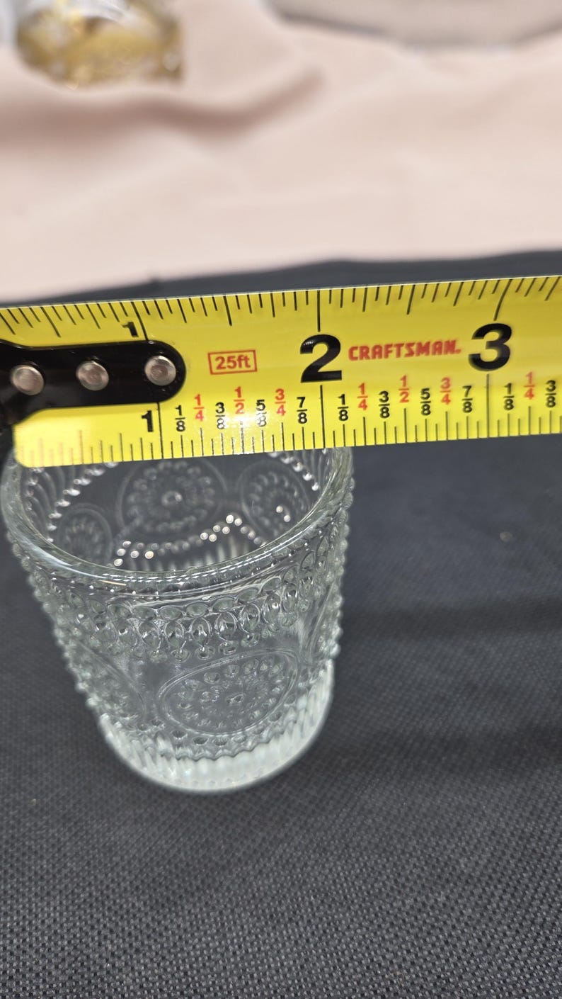 May include: A clear glass votive candle holder with a decorative pattern is measured with a yellow Craftsman tape measure. The tape measure reads approximately 2 inches across the top of the glass. The candle holder is on a dark surface.