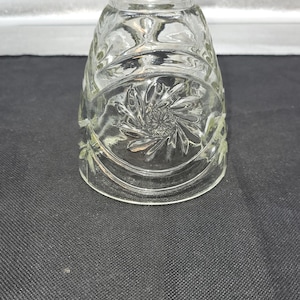 May include: A clear glass lampshade with a floral design. The lampshade has a flared shape and is decorated with embossed flowers and leaves. The glass is transparent, allowing light to pass through. The lampshade is likely for a table lamp or ceiling fixture.