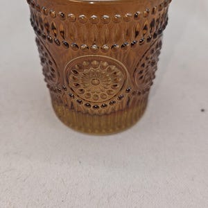 May include: A brown glass votive candle holder with a detailed, embossed design. The glass has a warm amber hue and features a repeating pattern of small circles and floral medallions. The rim has a series of small, raised bumps.