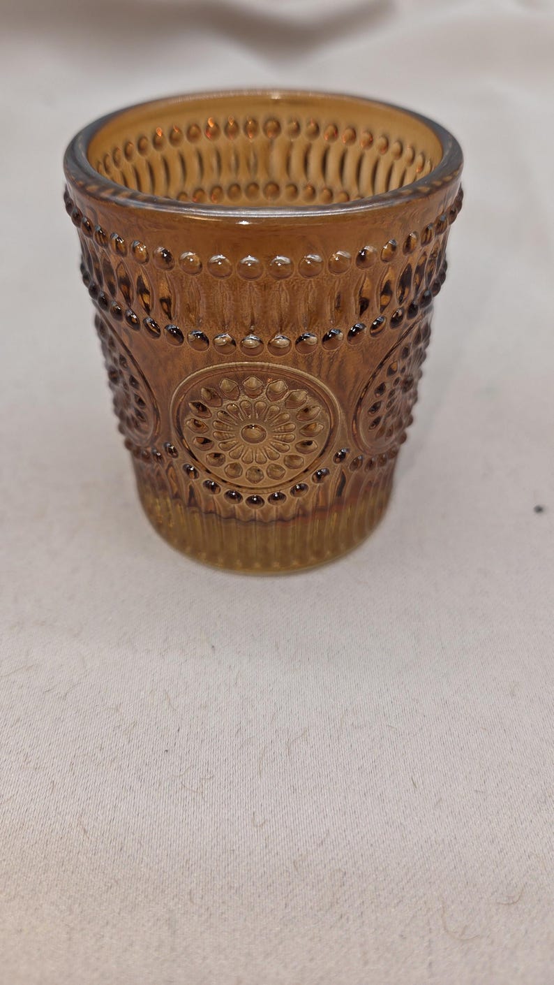 May include: A brown glass votive candle holder with a detailed, embossed design. The glass has a warm amber hue and features a repeating pattern of small circles and floral medallions. The rim has a series of small, raised bumps.