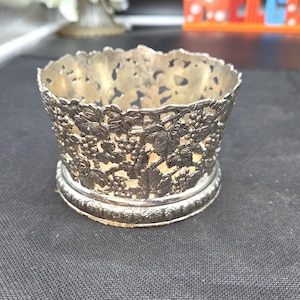 May include: A silver-toned metal coaster or bottle holder featuring an intricate openwork design of grapevines, leaves, and clusters of grapes. The circular object has a scalloped top edge and a beaded base rim with a metallic finish.