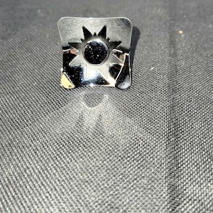 May include: A square, silver-colored metal ring with a sunburst design. The ring features a central circular element surrounded by pointed rays. The ring is set against a dark, textured background.