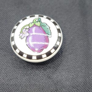 Puede incluir: Un pomo de cerámica blanco con un borde blanco y negro. El pomo presenta una berenjena morada con hojas verdes y una flor morada.