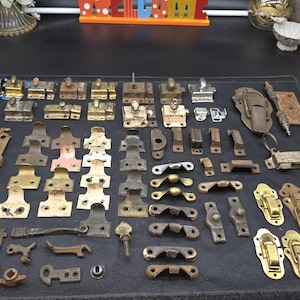 May include: A collection of assorted vintage metal hardware including brass and iron latches, window lifts, sash locks, hinges, and fasteners arranged in rows. The items feature various metallic finishes, including polished brass, aged iron, and weathered steel.