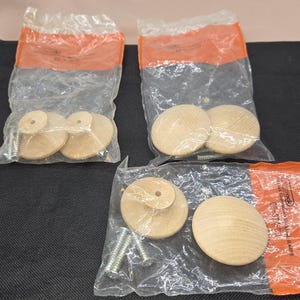 May include: Several packages of unfinished wooden cabinet knobs with screws. The knobs are round with a flat top and a smaller, raised circular section in the center. The knobs are packaged in clear plastic bags with orange labels.