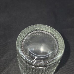 May include: Clear glass bottle with a textured, ridged design. The bottle has a wide, flat base and a slightly curved body. The glass is transparent, allowing light to pass through. The bottle is likely for storing liquids.