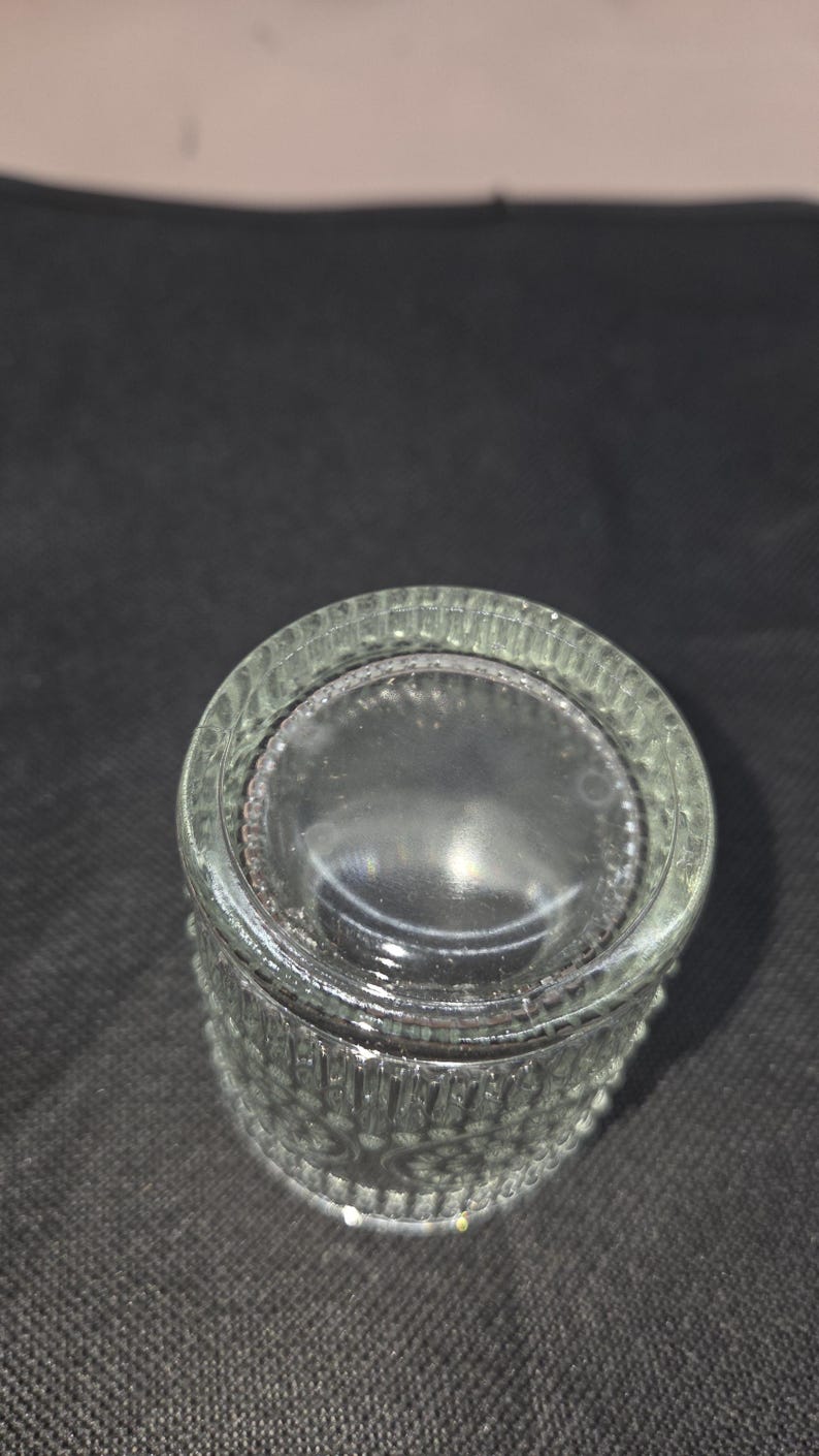 May include: Clear glass bottle with a textured, ridged design. The bottle has a wide, flat base and a slightly curved body. The glass is transparent, allowing light to pass through. The bottle is likely for storing liquids.
