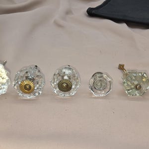 May include: A collection of six decorative cabinet knobs. The knobs vary in shape and design, including faceted crystal, clear glass with brass accents, and a gold-toned woven style. They are arranged on a light pink surface.