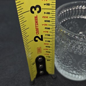 May include: A clear glass votive candle holder with a decorative pattern, measuring approximately 2 inches (5 cm) in height. A yellow Craftsman tape measure is positioned next to the glass, showing the measurement.