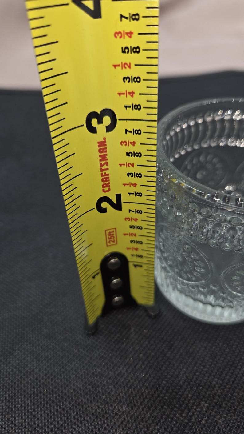 May include: A clear glass votive candle holder with a decorative pattern, measuring approximately 2 inches (5 cm) in height. A yellow Craftsman tape measure is positioned next to the glass, showing the measurement.
