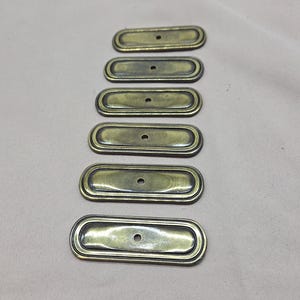 May include: Six antique brass-toned drawer pulls arranged vertically. Each pull is rectangular with rounded ends, a raised border, and a single centered hole. The hardware has a vintage aesthetic.