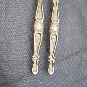 May include: Two elongated metal drawer pulls with a dark brass finish. Each handle features a symmetrical, ornate design with a textured central panel and smooth, curved edges that taper toward the top and bottom ends of the hardware piece.