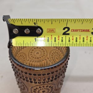 May include: Amber-colored glass votive candle holder with a detailed pattern. The votive is approximately 1.75 inches in diameter. The design features a series of raised dots and circular patterns. A yellow measuring tape is placed next to the votive.