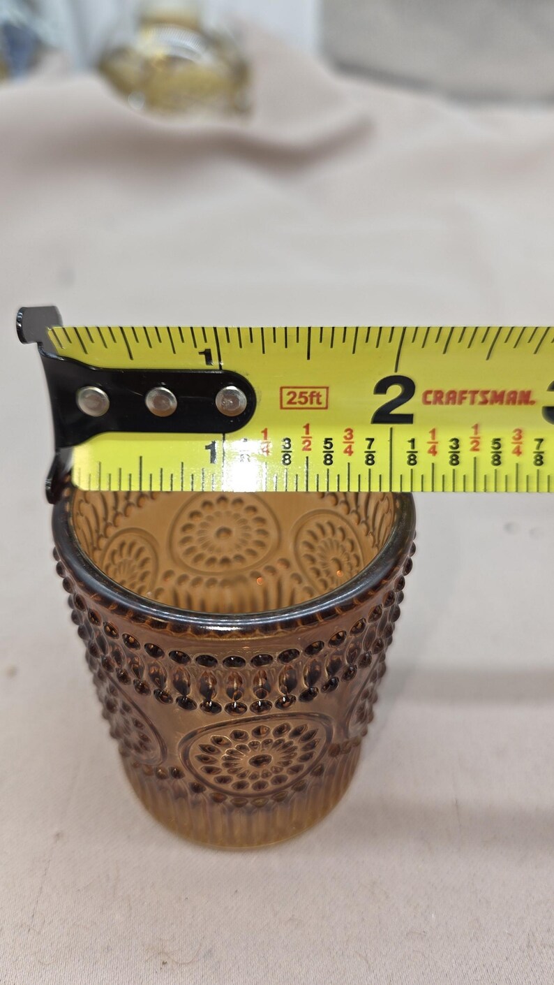 May include: Amber-colored glass votive candle holder with a detailed pattern. The votive is approximately 1.75 inches in diameter. The design features a series of raised dots and circular patterns. A yellow measuring tape is placed next to the votive.