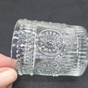 May include: A clear glass votive candle holder with a detailed, embossed design. The design includes circular patterns and beaded borders. The candle holder is cylindrical and appears to be made of thick glass.
