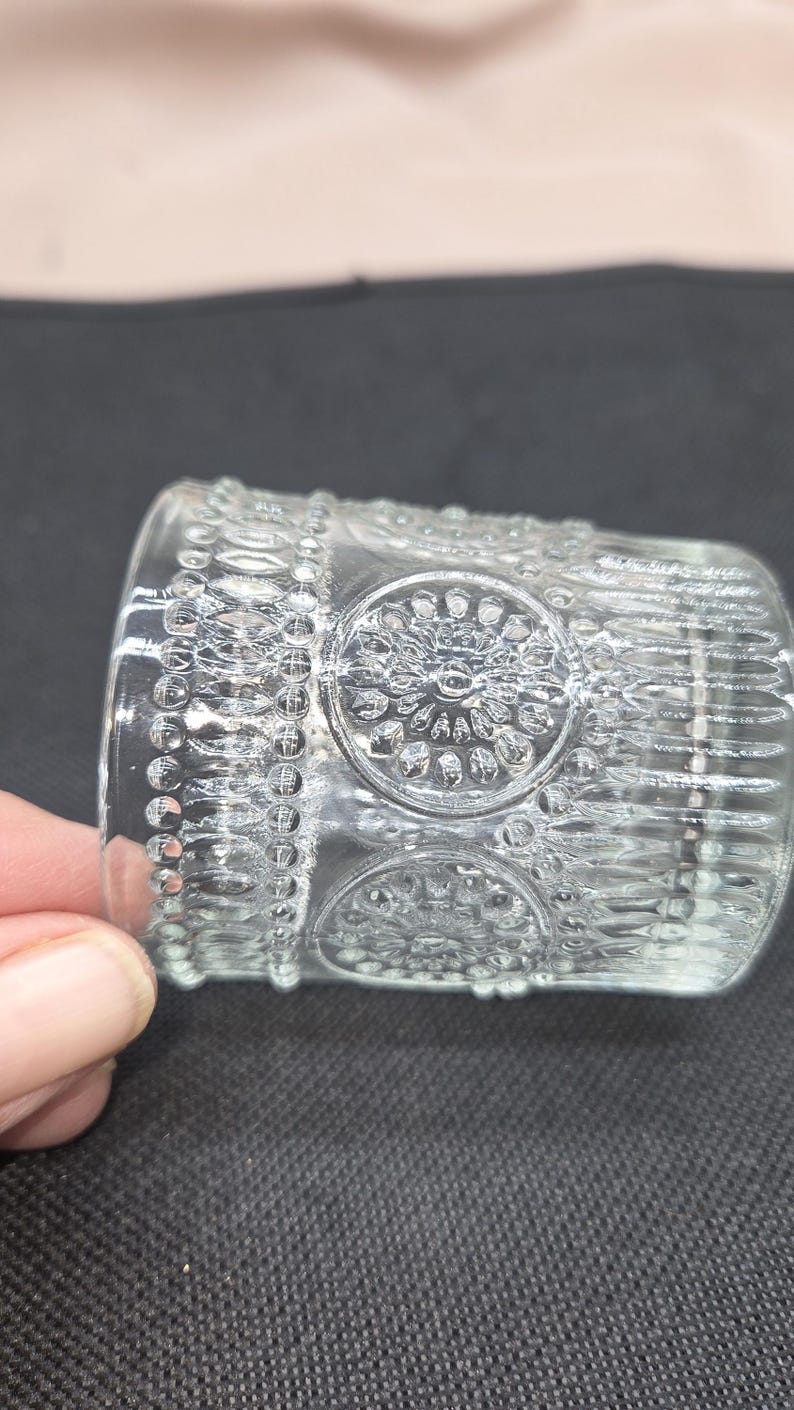 May include: A clear glass votive candle holder with a detailed, embossed design. The design includes circular patterns and beaded borders. The candle holder is cylindrical and appears to be made of thick glass.