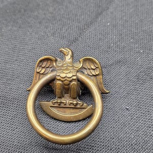 May include: A brass-toned metal drawer pull featuring a detailed eagle figure with wings spread, perched atop a semi-circular base. A circular ring hangs from the eagle, providing a functional handle for furniture or cabinetry hardware.