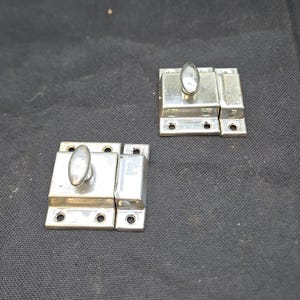 May include: Two silver-colored metal cabinet latches, each featuring a rectangular base plate with four screw holes and a central oval-shaped rotating knob mechanism designed to secure a door or drawer in a closed position.