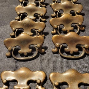 May include: Ten antique brass cabinet drawer pulls with ornate designs. The pulls have a loop handle and are in good condition.