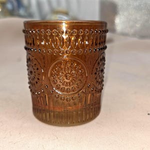 May include: A decorative amber-colored glass votive candle holder. The glass features an embossed design of circles and floral patterns. The candle holder is cylindrical and has a beaded rim. The overall theme is home decor.