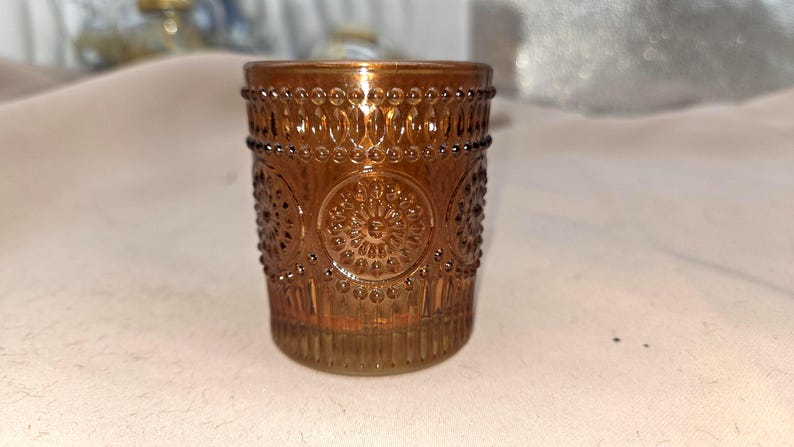 May include: A decorative amber-colored glass votive candle holder. The glass features an embossed design of circles and floral patterns. The candle holder is cylindrical and has a beaded rim. The overall theme is home decor.