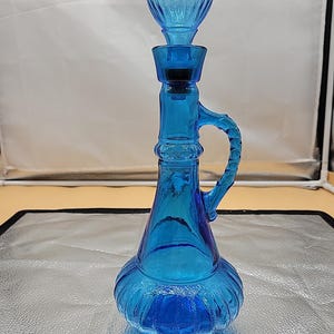 May include: A blue glass decanter with a wide, round body and a tall, slender neck. The decanter has a single handle and a removable stopper.