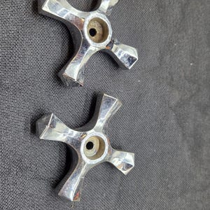 May include: Two chrome-plated metal faucet handles with a four-pointed cross design. Each handle features a central circular opening with a visible screw hole and faceted edges that taper toward the ends of each arm, reflecting light on the surface.