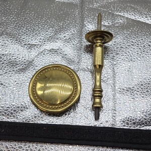 May include: A set of two brass-colored hardware pieces. One is a round, flat plate with a beaded edge. The other is a decorative screw with a detailed, turned design. The hardware is likely for furniture or decorative use.