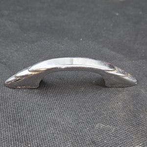 May include: A polished chrome cabinet handle with a curved, arching design. The hardware features a smooth, reflective metallic finish with tapered ends and two mounting points on the underside, showcasing a sleek, solid construction throughout.