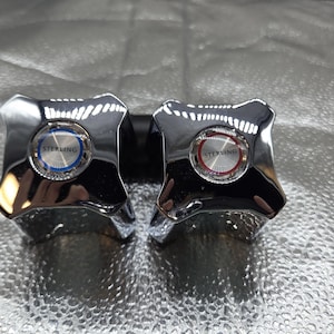 May include: Two chrome faucet handles with a four-point star design. Each handle has a circular center with the word "STERLING" and a blue or red accent. The handles are mounted on a black base.