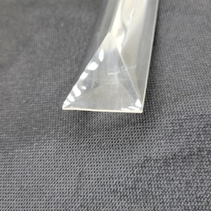 May include: A clear, triangular prism with a polished surface. The prism is curved and appears to be made of glass or a similar transparent material. The object is set against a dark gray textured background.
