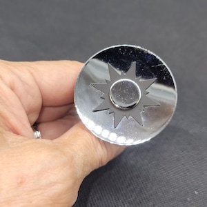 May include: A shiny, silver bottle stopper with a sunburst design. The stopper is round with a central circular detail and a starburst pattern around it. The stopper is held in a hand.