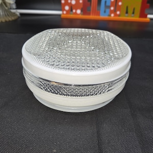 May include: A round, clear glass light fixture with a textured surface and a white rim. The fixture is sitting on a dark surface. In the background, a colorful "WELCOME" sign is visible.